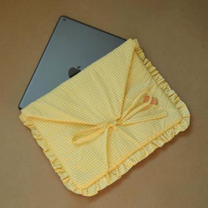 May include: A yellow and white gingham fabric tablet sleeve featuring a triangular flap closure with fabric ties and a ruffled border. A dark gray tablet with a visible Apple logo is partially inserted into the protective fabric case.