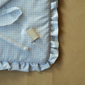May include: A light blue and white gingham patterned fabric cushion with a ruffled edge and fabric ties. A small, rectangular, off-white tag is sewn onto the surface with the word PLON embossed in capital letters on the front of the tag.