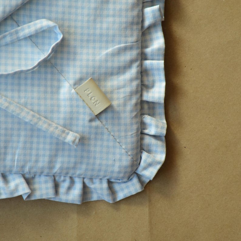 May include: A light blue and white gingham patterned fabric cushion with a ruffled edge and fabric ties. A small, rectangular, off-white tag is sewn onto the surface with the word PLON embossed in capital letters on the front of the tag.