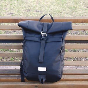 May include: A black roll-top backpack with a silver buckle and a small white label. The backpack has a top handle and adjustable shoulder straps. It is on a wooden bench.