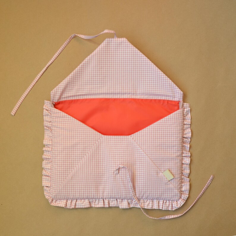 May include: A fabric envelope-style pouch featuring a light pink and white gingham pattern. The exterior is trimmed with a ruffled edge, and the interior is lined with a solid coral-colored fabric. Two thin fabric ties are attached for closure.