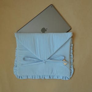 Pode incluir: Uma capa de tecido para portátil em azul claro com aba triangular estilo envelope, fecho de laço e bordas com folhos nas laterais e na parte inferior. Um tablet cinzento com o logótipo da Apple está inserido na abertura superior.