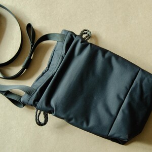 May include: A dark blue drawstring bag with a long adjustable strap. The bag is made of a water-resistant material and has a simple, minimalist design.