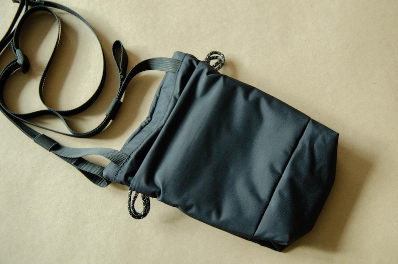 May include: A dark blue drawstring bag with a long adjustable strap. The bag is made of a water-resistant material and has a simple, minimalist design.