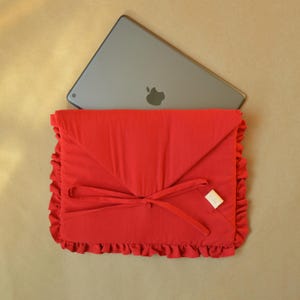 Op de afbeelding: Een felrode tablethoes met een gerimpelde rand en een striksluiting. De hoes is gemaakt van een zachte stof en heeft een klepsluiting. Een zilveren tablet met een Apple-logo is gedeeltelijk zichtbaar.