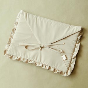 May include: A beige baby changing mat in an envelope shape, featuring a ruffled edge and a bow tie closure. The mat is made of a soft, quilted material. A small tag is attached. The design is simple and elegant.