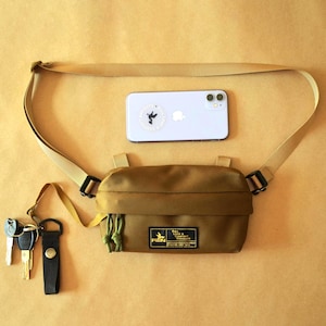 May include: A tan crossbody bag with an adjustable strap, featuring a rectangular black patch with yellow text reading PION PULL LOAD & COMFORT NAVIGATE. A light purple smartphone with a circular sticker and a set of keys on a black fob are included.