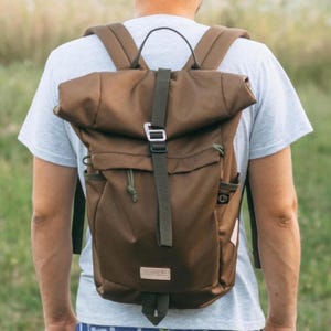 Op de afbeelding: Een bruine roll-top rugzak met een donkergroene riem en gespsluiting. De rugzak heeft twee zijzakken en een kleine patch aan de voorkant. De riemen zijn verstelbaar.