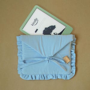 Könnte beinhalten: Eine hellblaue Stoffhülle mit gerüschtem Rand und Bindeband, versehen mit einem kleinen rechteckigen beigen Etikett mit der Aufschrift FLOR. Ein hellgrüner Kindle E-Reader mit weißem Bildschirm und Kindle-Logo ist darin eingesteckt.