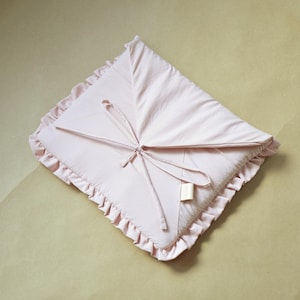 Könnte beinhalten: Eine hellrosa Babydecke mit gerüschtem Rand und Bindeband. Die Decke ist quadratisch gefaltet und hat ein kleines angebrachtes Etikett. Der weiche Stoff ist einfarbig.