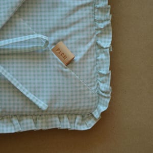 May include: A light mint green and white gingham patterned fabric item featuring a ruffled edge and a small rectangular tan leather tag embossed with the letters PLCN. The textile includes a thin fabric tie attached to the corner of the piece.