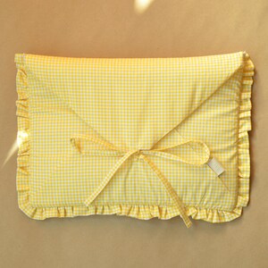 May include: A rectangular fabric pouch featuring a yellow and white gingham pattern. The envelope-style closure is secured with two fabric ties, and the entire perimeter of the pouch is finished with a gathered ruffle trim in the same fabric.