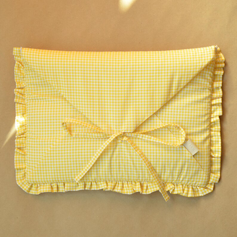 May include: A rectangular fabric pouch featuring a yellow and white gingham pattern. The envelope-style closure is secured with two fabric ties, and the entire perimeter of the pouch is finished with a gathered ruffle trim in the same fabric.