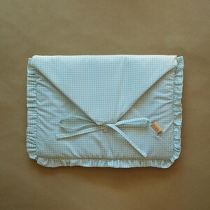 May include: A rectangular fabric pouch featuring a light mint green and white gingham pattern. The envelope-style flap is secured with matching fabric ties. The edges are finished with a ruffled trim, and a small tan label reads FLON.