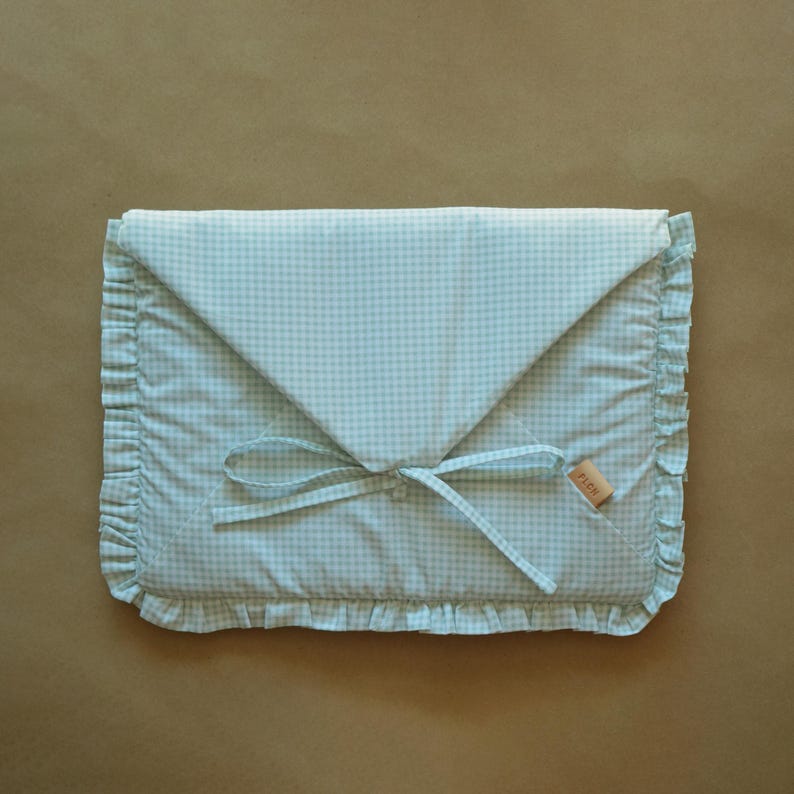 May include: A rectangular fabric pouch featuring a light mint green and white gingham pattern. The envelope-style flap is secured with matching fabric ties. The edges are finished with a ruffled trim, and a small tan label reads FLON.