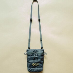 May include: A dark gray crossbody bag with a zippered pocket and a small logo patch. The bag has a long adjustable strap.