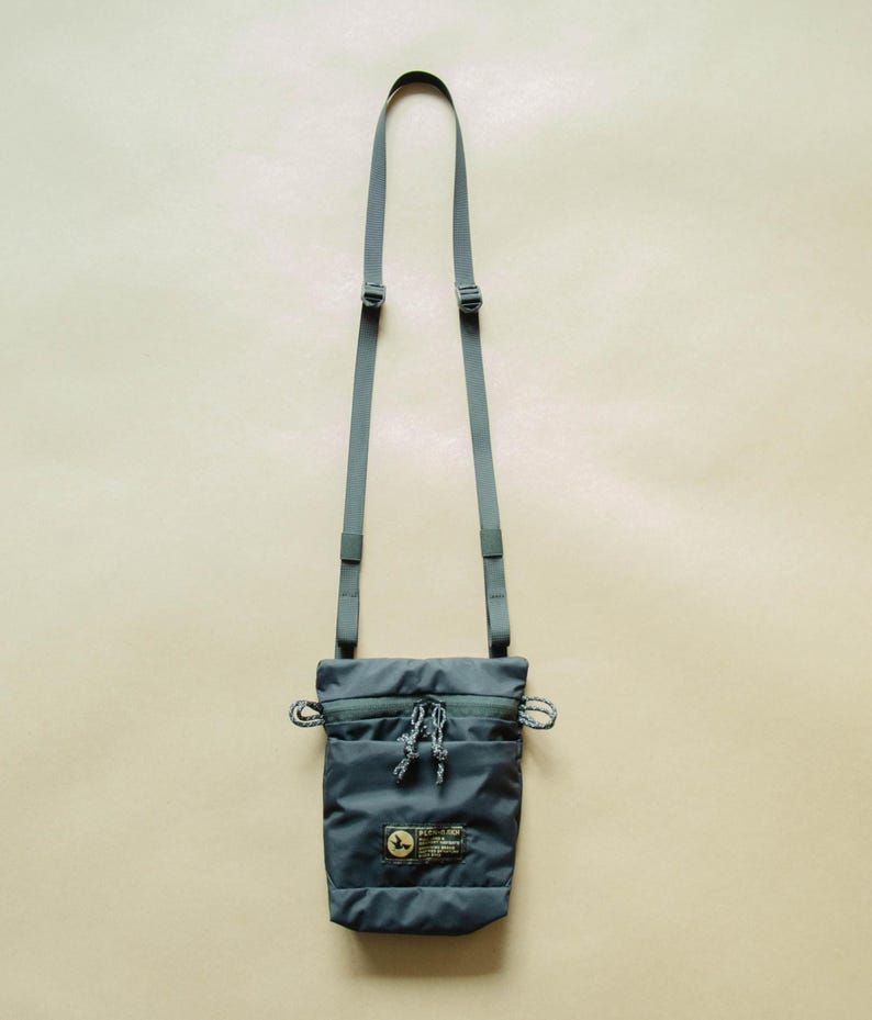 May include: A dark gray crossbody bag with a zippered pocket and a small logo patch. The bag has a long adjustable strap.