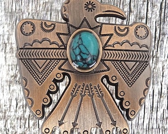 May include: A bronze-colored metal pin with a turquoise stone in the center, depicting a stylized Native American Thunderbird with arrows.
