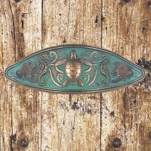 Op de afbeelding: Een ovaalvormig decoratief stuk in teal kleur met een gouden zeeschildpad, zeepaardjes en schelpen. Het stuk is gemaakt van metaal en heeft een getextureerd oppervlak.