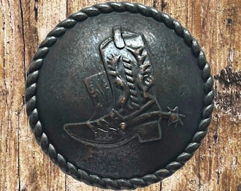 Puede incluir: Un botón redondo de metal marrón oscuro con un diseño en relieve de una bota de vaquero con espuela. El botón tiene un borde de cuerda.