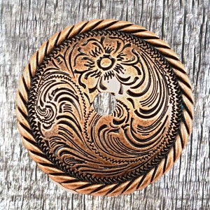 May include: A copper-colored metal knob with a decorative floral and swirl design. The knob has a rope-like border and a hole in the center.