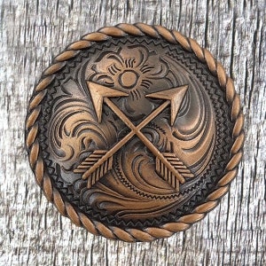 以下が含まれることがあります： ロープ状の縁取りと渦巻く花柄が施された銅色のベルトバックル。バックルの中央には、交差した矢印が2本あります。