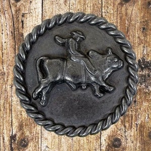 May include: A silver-toned metal belt buckle with a rope design border. The buckle features a cowboy riding a bull.