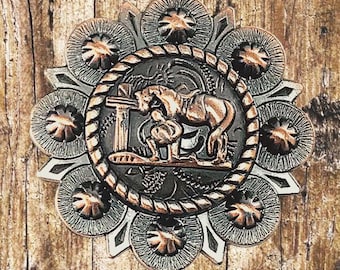 Puede incluir: Un concho metálico circular detallado con temática del Oeste. El centro presenta un relieve de un caballo y un jinete, rodeado por un borde en forma de cuerda. El anillo exterior tiene un diseño floral con acabado plateado y cobrizo. Sobre fondo de madera.