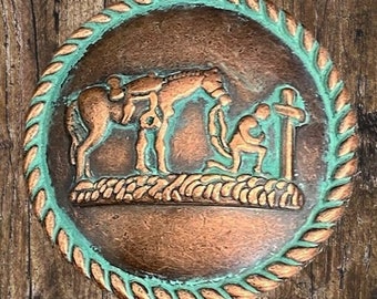 Puede incluir: Una placa redonda de color cobre con un borde de cuerda en color verde turquesa. La placa presenta una escultura en relieve de un caballo arrodillado con la cabeza inclinada, una persona arrodillada en oración y una cruz.