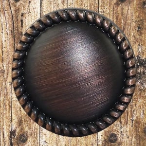 Peut inclure: Un bouton rond en métal brun foncé avec un motif de corde autour du bord. Le bouton a une finition brossée.