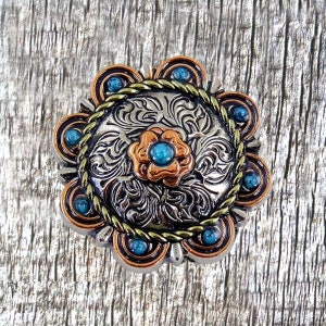 May include: A silver and copper-toned metal belt buckle with a floral design and turquoise stones. The buckle has a rope-like border and is surrounded by a circle of turquoise stones.