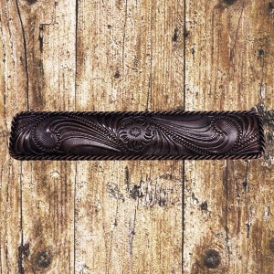 Pode incluir: Uma puxador de gaveta de metal marrom escuro com uma borda de corda e um design floral intrincado. O puxador está sobre um fundo de madeira.