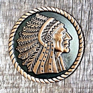 May include: A copper-colored metal pin with a detailed image of a Native American chief wearing a headdress. The pin is circular and has a rope-like border.