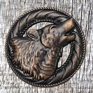 Puede incluir: Un botón de metal de color bronce con un diseño detallado de la cabeza de un lobo aullando a la luna. El lobo está rodeado por un borde de cuerda.