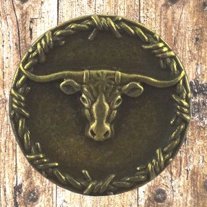 Peut inclure: Un bouton en métal de couleur bronze avec une bordure de fil de fer barbelé et une image en relief d'une tête de taureau à longues cornes au centre.