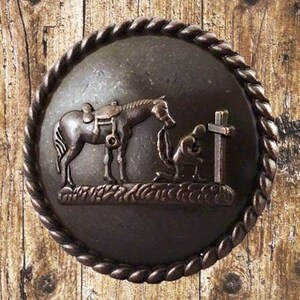 Peut inclure: Une boucle de ceinture en métal de couleur bronze avec une bordure en forme de corde. La boucle présente une sculpture en relief d'un cheval avec une selle et une personne agenouillée devant une croix.