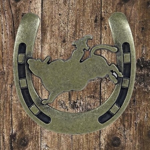 Peut inclure: Un fer à cheval en métal rustique avec une silhouette de cowboy chevauchant un taureau en train de s'emballer. Le fer à cheval est de couleur bronze foncé avec des accents noirs.