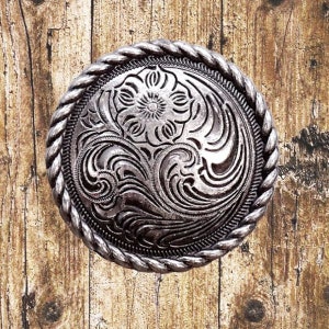 May include: A silver-toned metal belt buckle with a rope-like border and an intricate floral design.