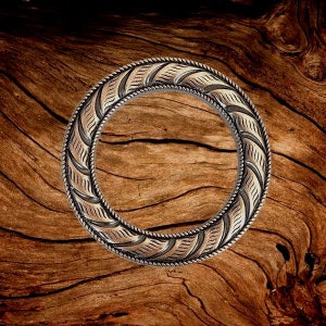 May include: A silver-toned metal ring with a decorative design of leaves and a rope-like border. The ring is on a wooden surface.