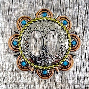 May include: Silver and copper buckle with a turquoise and gold design. The buckle has a rope-like border and a floral pattern.