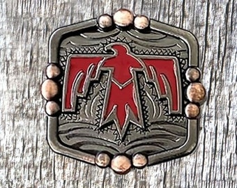 May include: A silver-toned metal belt buckle with a red stylized bird design. The buckle has a hexagonal shape with rounded edges and a decorative border. The bird is centered in the buckle and has a detailed design with intricate lines and patterns.
