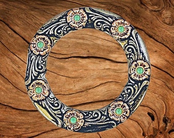 May include: A silver ring with a decorative design featuring green and gold flowers. The ring is on a wooden background.