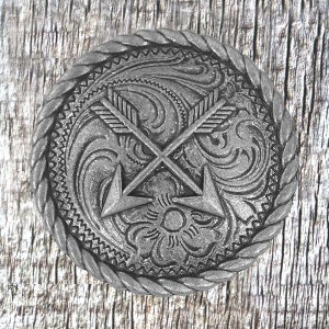 以下が含まれることがあります： ロープ状の縁取りと、2本の交差した矢印と花の模様の装飾的なデザインが施された銀色の金属製のベルトバックルです。