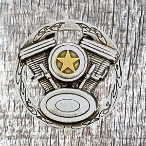 Può includere: Fibbia per cintura in metallo argentato con un design dettagliato di un motore di motocicletta. Il motore ha una stella dorata al centro e un cerchio argentato intorno. La fibbia è circondata da un design a fiamma argentato.
