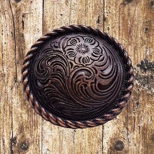 以下が含まれることがあります： ロープ状の縁取りと複雑な花柄が施された、丸い茶色の金属製のベルトバックルです。