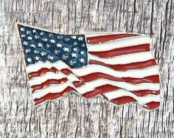 May include: A small, waving American flag pin with red, white, and blue enamel. The flag is on a wood background.