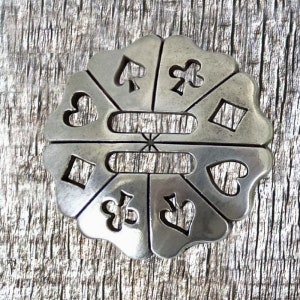 May include: A silver-toned metal charm with a circular design featuring cutouts of playing card suits: hearts, diamonds, spades, and clubs. The charm has a slightly textured surface.