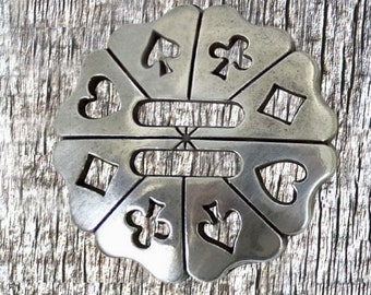May include: A silver-toned metal charm with a circular design featuring cutouts of playing card suits: hearts, diamonds, spades, and clubs. The charm has a slightly textured surface.