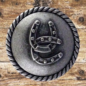 Puede incluir: Una hebilla de metal plateada en forma de herradura con un borde de cuerda. La herradura tiene un diseño de doble herradura.