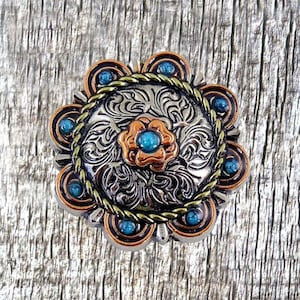 May include: A silver and copper-toned metal belt buckle with a floral design and turquoise stones. The buckle has a rope-like border and is surrounded by a circle of turquoise stones.